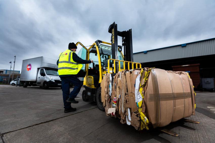 A man in a Mitie hi-vis vest climbing into a yellow forklift loaded with cardboard waste